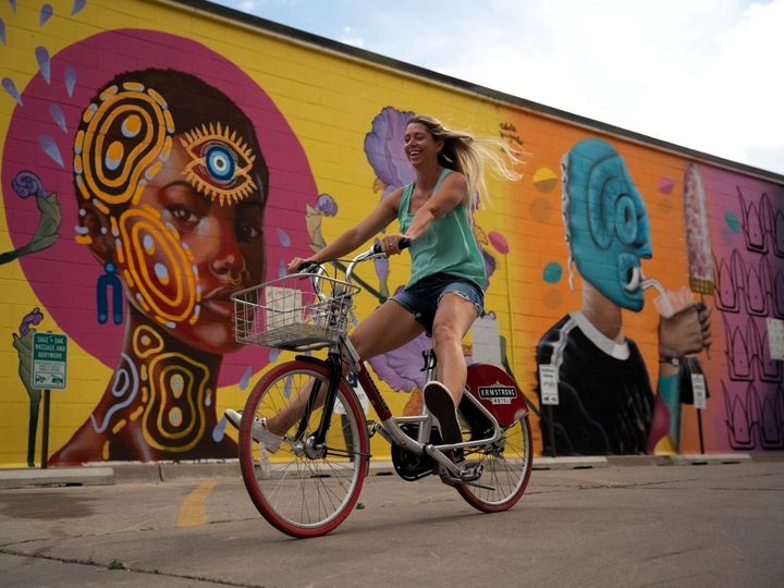 A woman rides a bike next to a colorful mural covering the entire exterior wall of a building.