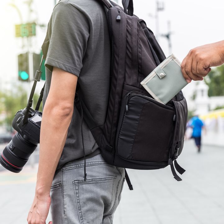 A pickpocket stealing a wallet out of the backpack of a tourist standing on the street carrying a camera.