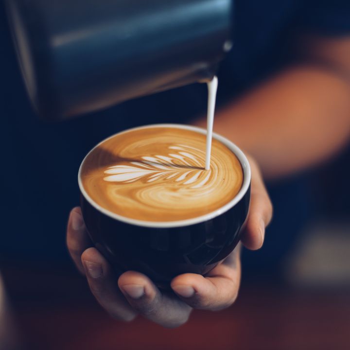 A hand is holding a cup with milk being artfully poured into a latte.