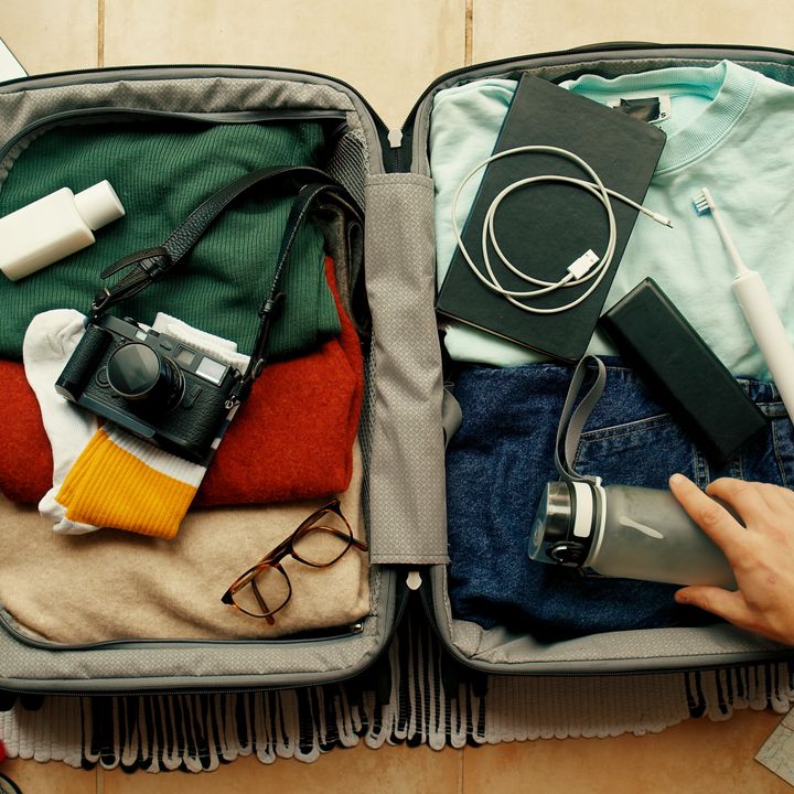 A hand is seen reaching into a suitcase packed full of clothes and accessories next to a laptop and other small items like headphones and a wallet and keyboard.