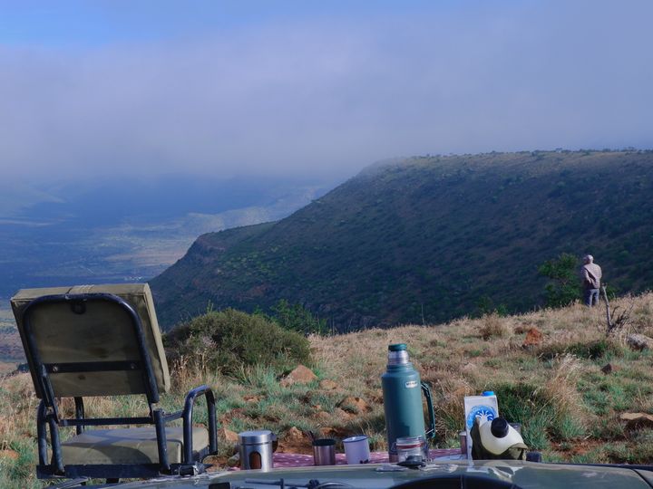 A picnic with a chair and a thermos and cups set up on blanket on a mountain.