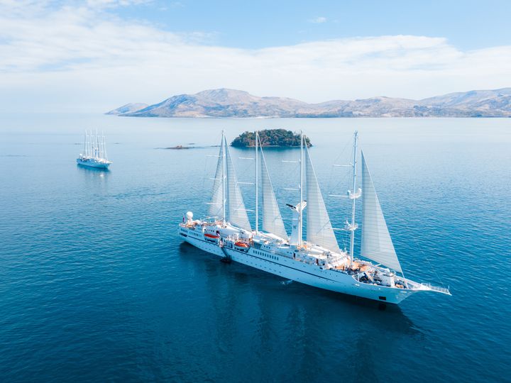 A tall sail ship from Windstar cruises is seen sailing not too far from land with a smaller ship behind it.