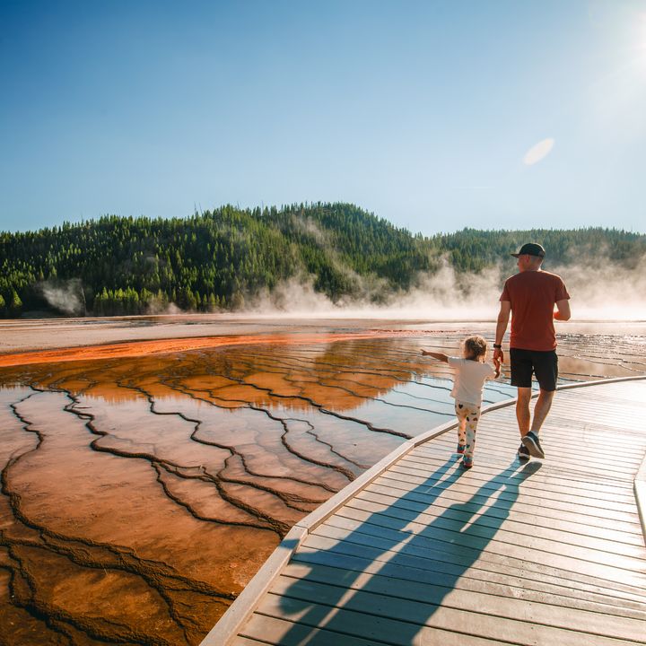 A man and his child walk on a boardwalk next to geysers at Yellowstone National Park.