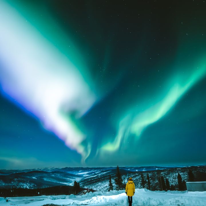 A person in a yellow coat stands on a snowy landscape in Fairbanks, Alaska as neon blue and green northern lights appear in the night sky.