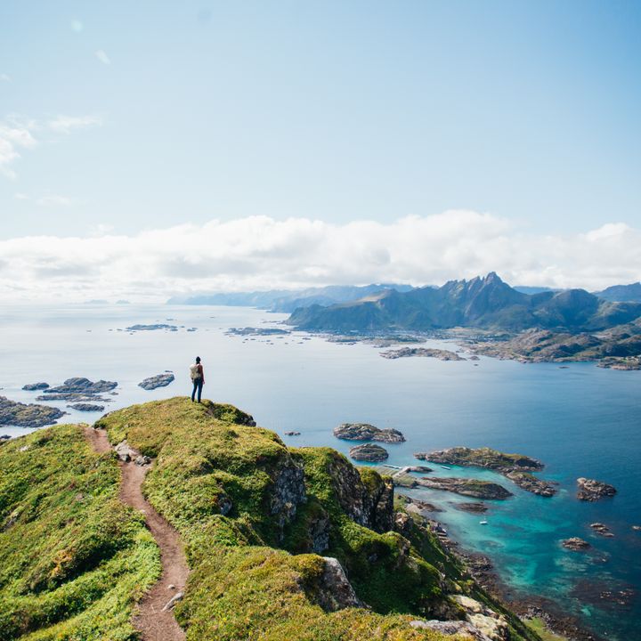 A hiker stands on top of a mountain after a hike in Lofoten Islands in Norway looking out over rocks, water, and mountains.