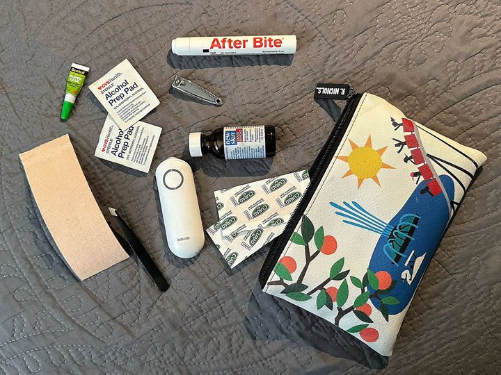 A travel pouch on a blanket surrounded by items such as nail clippers, alcohol pads, bug bite treatments, and super glue.