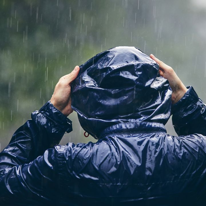 A person stands in the pouring rain, seen from behind, holding their hood up.