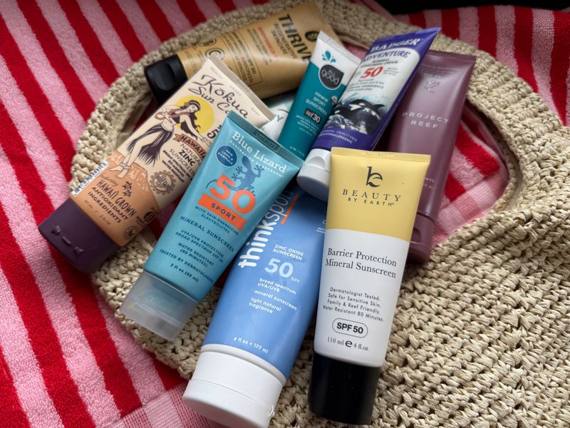 A collection of mineral sunscreen bottles arranged on top of a striped beach towel and a straw beach bag.