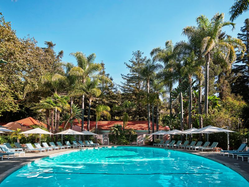 A round circular pool surrounded by pool chairs with a Mediterranean-style building behind it and towering green trees all around.