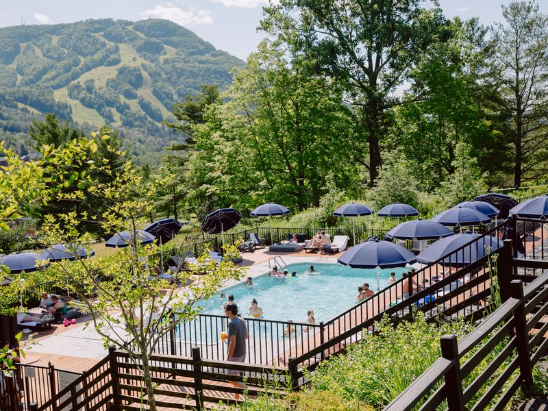 Stairs lead down to a pool surrounded by chairs and blue umbrellas next to green mountains.