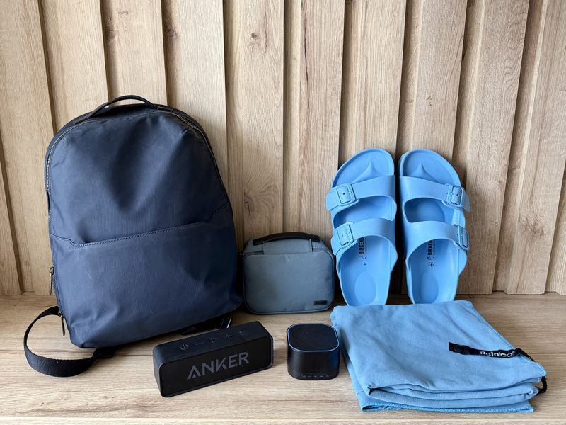 A backpack sits next to a travel pouch, a white noise machine, speaker, blue shoes, and a towel in front of a wood wall on wood floors.