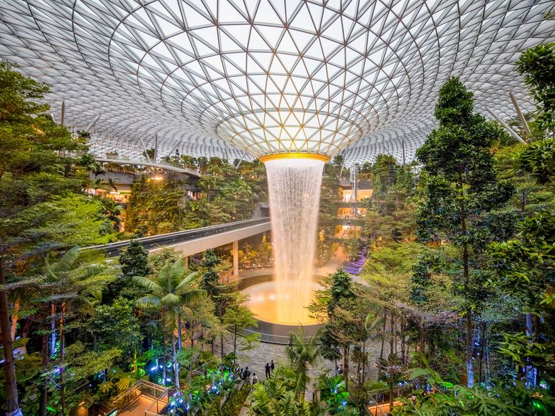 A waterfall cascades down from a structured ceiling surrounded by trees in a shopping area within Singapore’s Changi Airport.