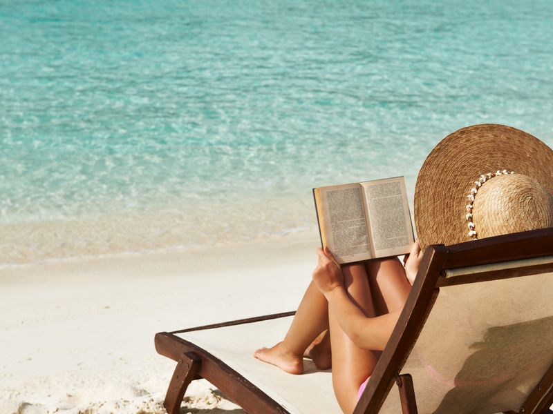 A woman is seen from behind reading a book while sitting on a beach chair facing crystal clean water and wearing a sun hat. 