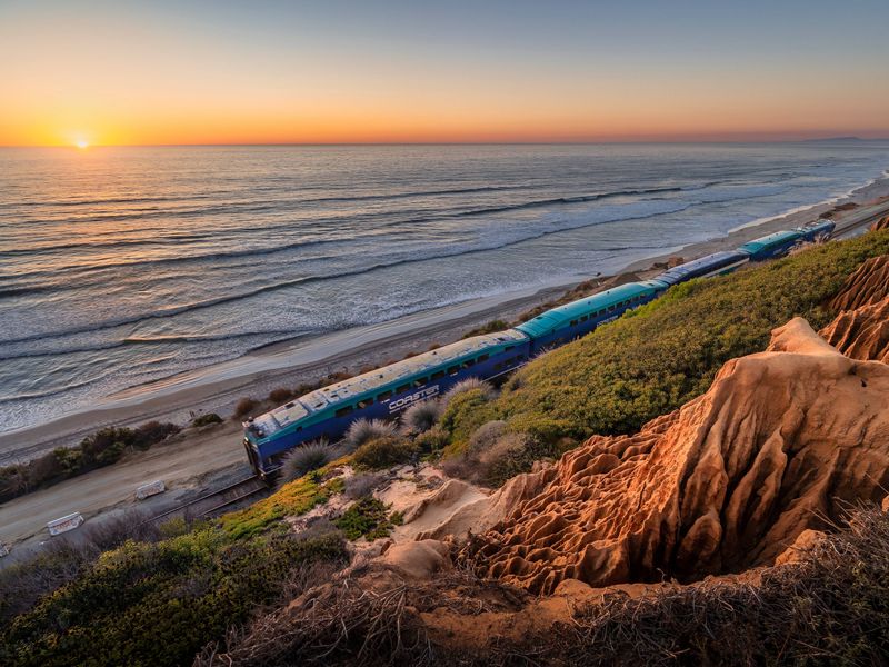 The Pacific Surfliner Amtrak train on a track in Del Mar, California that hugs the coastline and beach at sunset.